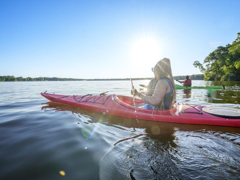 A man and a woman kayak on a large lake near the shore with trees on the edge of the land.